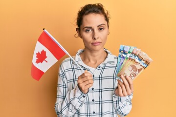 Poster - Young brunette woman holding canada flag and dollars relaxed with serious expression on face. simple and natural looking at the camera.