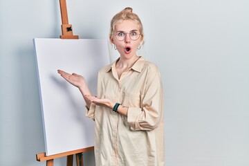 Poster - Beautiful caucasian woman with blond hair standing by painter easel stand presenting canva afraid and shocked with surprise and amazed expression, fear and excited face.