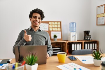 Canvas Print - Young hispanic man wearing business style sitting on desk at office doing happy thumbs up gesture with hand. approving expression looking at the camera showing success.