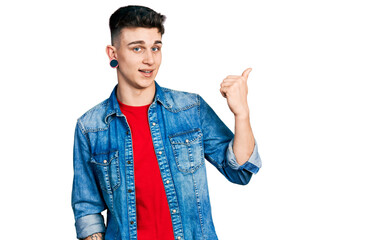 Young caucasian boy with ears dilation wearing casual denim jacket smiling with happy face looking and pointing to the side with thumb up.