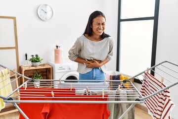 Poster - Young hispanic woman putting fresh laundry on clothesline with hand on stomach because indigestion, painful illness feeling unwell. ache concept.