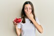 Young brunette woman holding red peppers covering mouth with hand, shocked and afraid for mistake. surprised expression