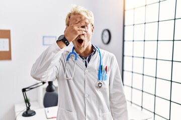 Sticker - Young blond man wearing doctor uniform and stethoscope at clinic peeking in shock covering face and eyes with hand, looking through fingers with embarrassed expression.