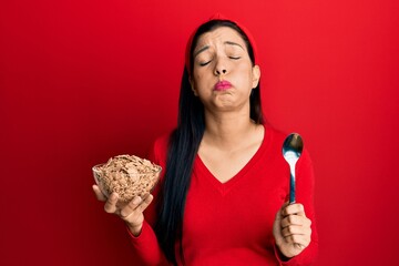 Sticker - Young latin woman holding cornflakes and spoon puffing cheeks with funny face. mouth inflated with air, catching air.