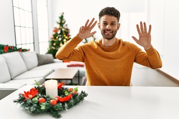 Sticker - Arab young man sitting on the table by christmas tree showing and pointing up with fingers number nine while smiling confident and happy.