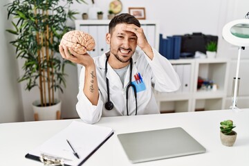 Wall Mural - Young doctor holding brain at medical clinic stressed and frustrated with hand on head, surprised and angry face