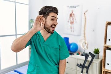 Poster - Young man with beard working at pain recovery clinic smiling with hand over ear listening an hearing to rumor or gossip. deafness concept.