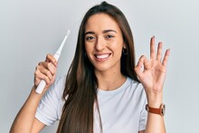 Young Hispanic Girl Holding Electric Toothbrush Doing Ok Sign With Fingers, Smiling Friendly Gesturing Excellent Symbol