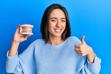 Wall Mural - Young hispanic girl holding orthodontic smiling happy and positive, thumb up doing excellent and approval sign