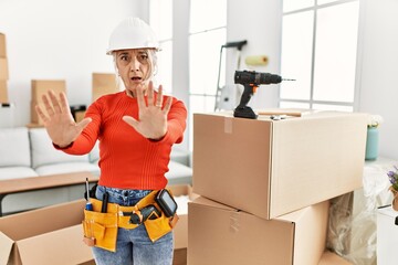 Canvas Print - Middle age grey-haired woman wearing hardhat standing at new home afraid and terrified with fear expression stop gesture with hands, shouting in shock. panic concept.