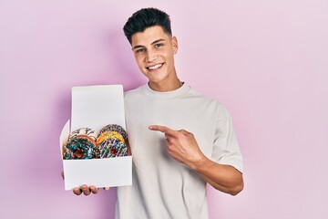 Canvas Print - Young hispanic man holding box of tasty colorful doughnuts smiling happy pointing with hand and finger