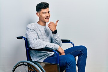 Canvas Print - Young hispanic man sitting on wheelchair smiling with happy face looking and pointing to the side with thumb up.