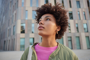 Wall Mural - Close up shot of thoughtful curly haired woman focused above thinks about something poses outdoors against modern building background walks in city during free time goes sightseeing explores place