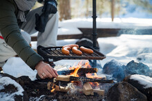 Man Roasting Sausages On Campfire In Forest By The Lake, Making A Fire, Grilling. Happy Tourist Exploring Finland. Beautiful Sunny Winter Landscape, Wood Covered With Snow. 