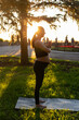 © satura_ - Side view of young woman expecting baby in black suit stands on rug barefoot and meditates on warm sunny summer day. Concept of sports and meditation activities