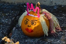 Orange Pumpkin Decorated As Princess With Painted Eyes And Mouth, Crown Flom Fluffy Pink Wire And Hair From Wooly Strings, Sunbathing In Afternoon Sunshine