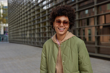 Wall Mural - outdoor shot of fashionable attractive woman smiles broadly wears sunglasses sweatshirt and jacket s