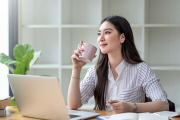 Beautiful young Asian businesswoman smiling holding a coffee mug and laptop working at the office.