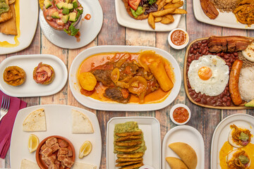 Top view image of a set of Colombian food dishes, with valluna cutlet, empanadas, paisa tray, prawn cocktail, fried patacones, shredded meat and guacamole