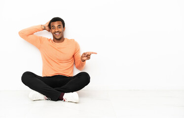 Wall Mural - African American man sitting on the floor over isolated copyspace background surprised and pointing finger to the side