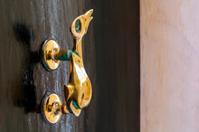 Closeup Of Brass Vintage Knocker As Dolphin With A Trident-shaped Tail At Black Wooden Door. Black Door With Cast Brass Hardware With Patina.