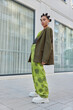 © wayhome.studio - Full body shot of millennial girl in stylish outfit white boots poses outside against some building looks at camera has serious expression leans behind. Female teenager with hair buns and makeup