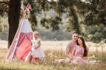 Wall Mural - Mom, dad and little daughter are sitting next to wigwam decoration in the park. Family spending time outdoor in summer, having fun together. Girl are dressed in pink dress.