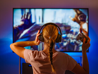 A girl shooter in headphones and with a joystick sits in front of a large monitor, plays a video game. Blue neon light, shooting from the back. Video games, strategy games, gambling, gamer, shooter.