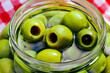 whole green pitted olives in liquid fill large clear glass jar closeup details with red and white gingham checked cloth napkin