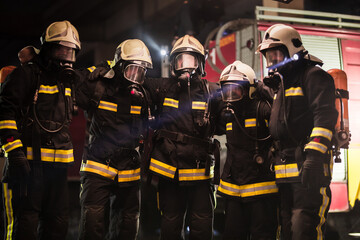 Wall Mural - Group of professional firefighters posing. Firemen wearing uniforms, protective helmets and oxygen masks. Smoke and firetrucks in the background.