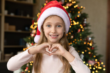 Head shot portrait of happy cute little 7s child girl in red santa hat showing heart sign with fingers, posing near decorated Christmas tree promoting support and help, miracle time, winter holidays.