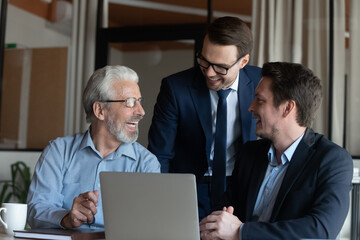 Wall Mural - Intergenerational business team. Smiling male colleagues of diverse age have fun by computer at workplace. Older and younger men office employees gather at laptop talk laugh joke on funny work moment