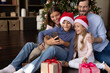 © fizkes - Happy bonding young couple parents and laughing little kids using cellphone, posing for funny selfie web camera photo, holding video call conversation, sitting on floor near decorated Christmas tree.