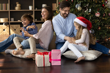 Happy couple parents having fun with small children sitting on pillows on floor in living room, enjoying playing together near decorated festive Christmas tree and wrapped boxes with presents gifts.