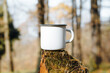 Camping metal white mug mock up standing on tree stump in woods outdoors. Enamelled balnk cup with empty space for branding or logo