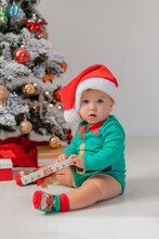Cute Baby In A Christmas Gnome Costume Opens Gifts Near The Christmas Tree. Products For Children And Holidays. Winter New Year's Concept. Space For Text. High Quality Photo