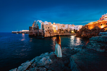 Wall Mural - Young woman on the rocks in front of the Apulian village of Polignano a Mare at sunset (blue hour)