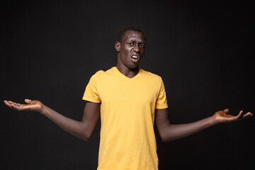 Wall Mural - Displeased perplexed young african american man guy in yellow t-shirt posing isolated on black wall background studio portrait. People emotions lifestyle concept. Mock up copy space. Spreading hands.