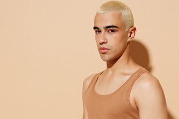 Side view young trendy serious blond latin hispanic gay man 20s with make up in beige tank shirt looking camera isolated on plain light ocher background studio portrait People lgbt lifestyle concept.