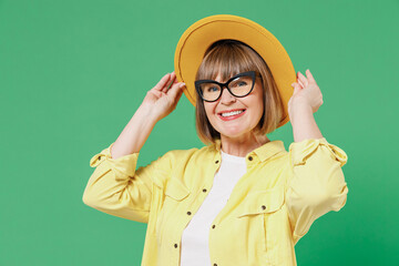 Elderly smiling happy fashionable woman 50s wearing glasses yellow shirt white t-shirt touch hat looking camera isolated on plain green background studio portrait People emotions lifestyle concept.