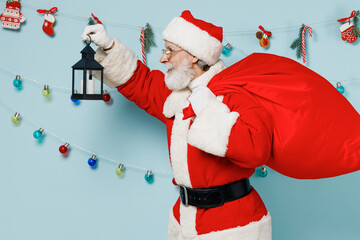 Old Santa Claus man 50s wears Christmas hat red suit clothes carrying bag with gifts lantern isolated on plain blue background studio. Happy New Year 2022 celebration merry ho x-mas holiday concept.