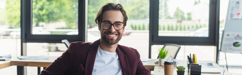 Wall Mural - Smiling businessman in eyeglasses looking at camera in office, banner