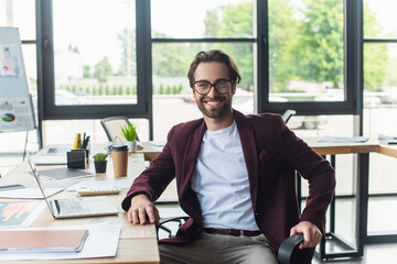 Wall Mural - Positive businessman in eyeglasses looking at camera near devices, calculator and papers in office