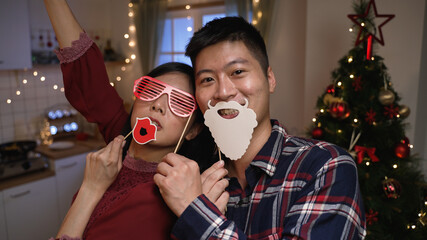 Wall Mural - funny asian couple looking at mobile cam is playing with xmas masks on stick while they are having fun taking selfies with Christmas tree on background at home