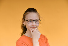 A Young Girl In Glasses Holds Her Face With Her Hand And Looks Thoughtfully Into The Camera. Portrait Of A Girl On A Yellow Background.