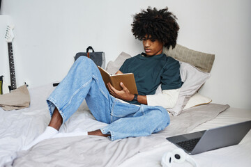 Wall Mural - Full length portrait of young African-American man sitting on bed at home and writing in notebook, copy space