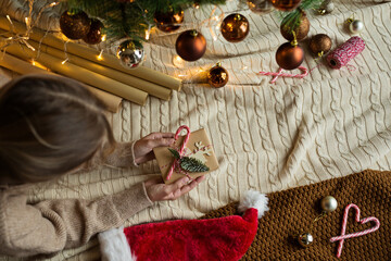 Wall Mural - Christmas composition. Woman wrapping gift box in craft paper near fir tree, next lying santa hat, candy canes. new year concept. Xmas celebration 2022. Flat lay, top view, overhead