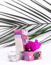 Creative Tropical Concept Made Of Organic Natural Soap Bars With Leaves And Rock On White Background With Palm Tree Branch Standing Behind Soaps. 
