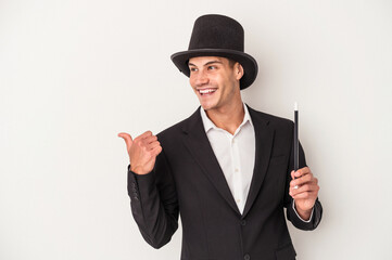 Young magician caucasian man holding a wand isolated on white background points with thumb finger away, laughing and carefree.