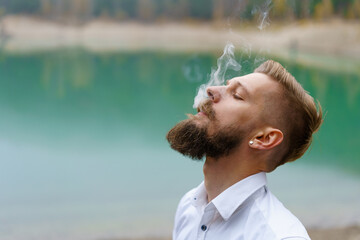 Business caucasian young bearded man in a white shirt smokes a cigarette and blows smoke against the backdrop of a turquoise lake in reflections. Faces solving problems and making choices